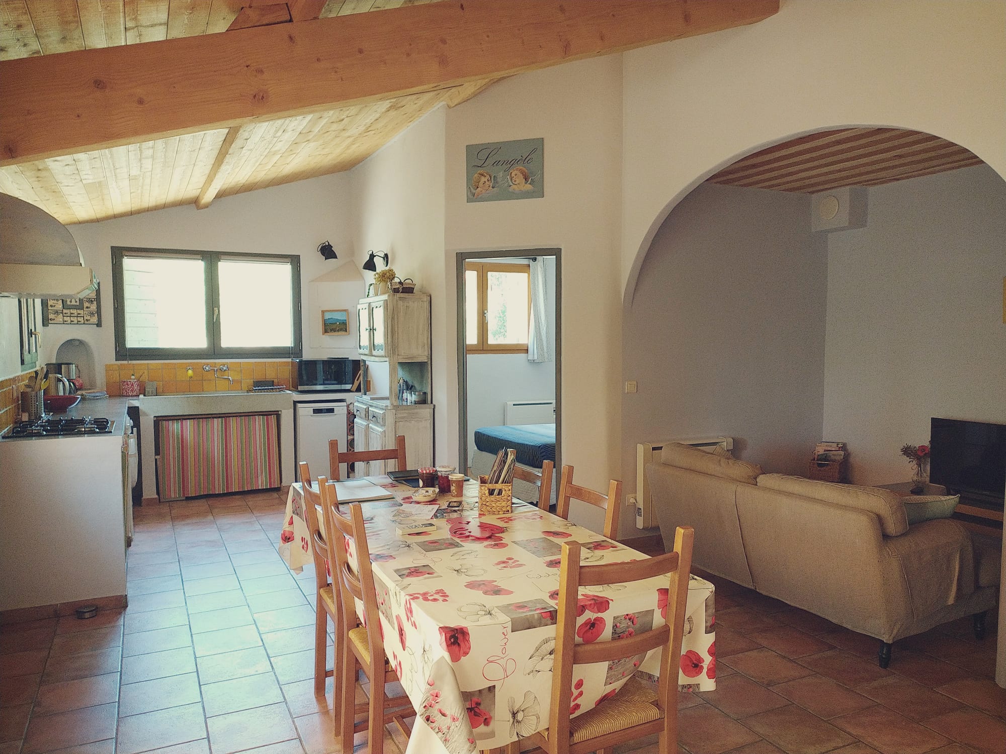 Cozy kitchen and living area with wooden beams, tiled floors, a floral tablecloth, and soft beige furniture. Bright and inviting space.